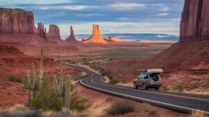 A winding road cuts through the Desert Southwest, passing red rock formations and mesas. An SUV with a roof box heads toward distant cliffs. Saguaro cacti dot the scene beneath soft clouds, highlighting Monument Valley's iconic road trip landscape.